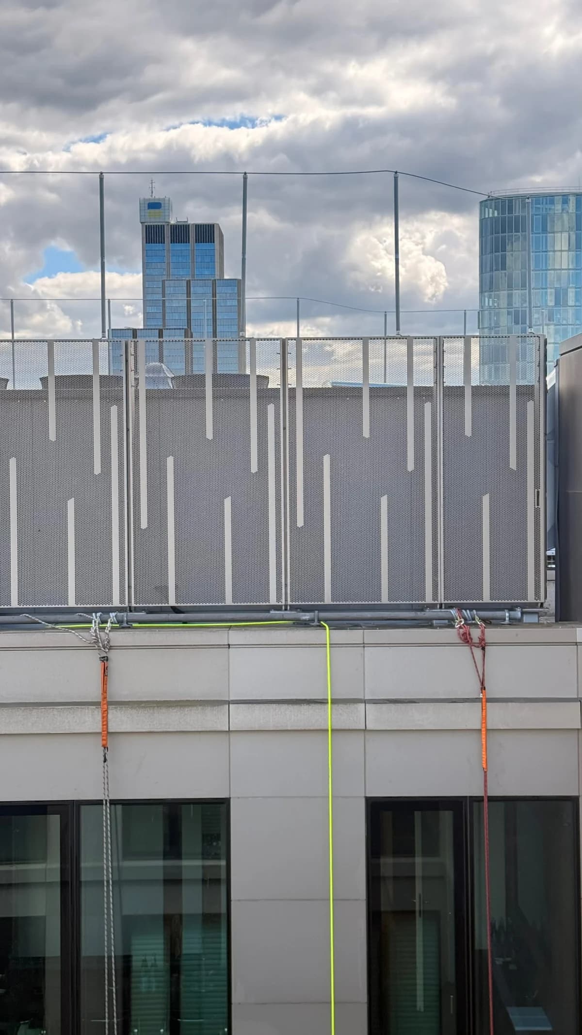 Metallverkleidung, Dachkante und obere Fenster einer Fassade mit darüber verlaufenden Seilen.